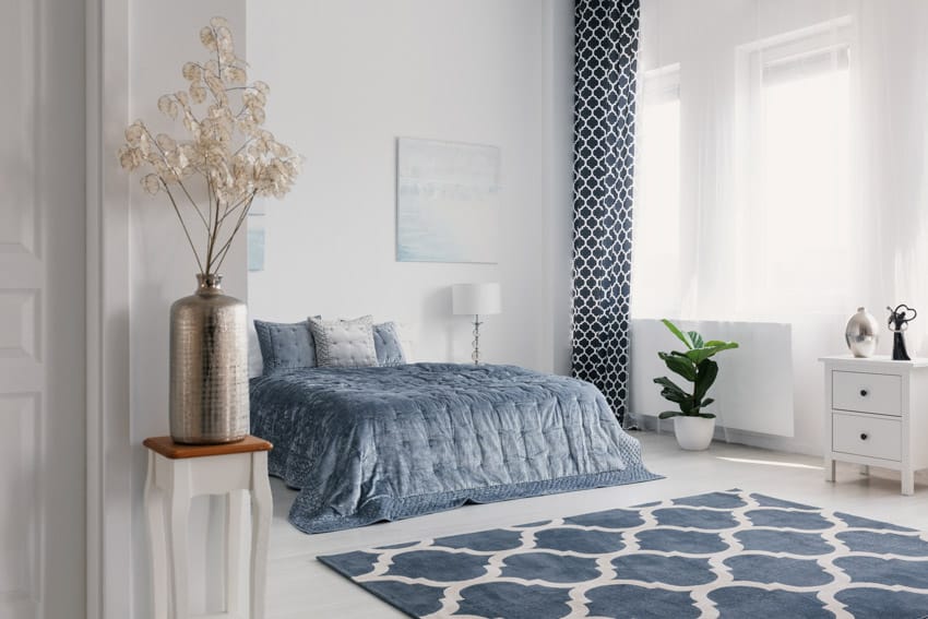 Bedroom with blue comforter, lamp, floor rug, indoor plant, decor pieces, and window curtain