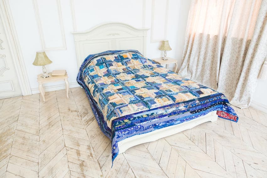 Bedroom with herringbone pattern wood floor, comforter, nightstands, headboard, window curtain, and white wall