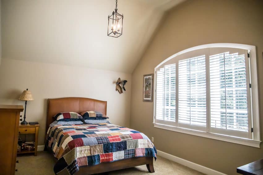 Bedroom with patterned comforter, wood headboard, pendant light, window, nightstand, lamp, and beige walls