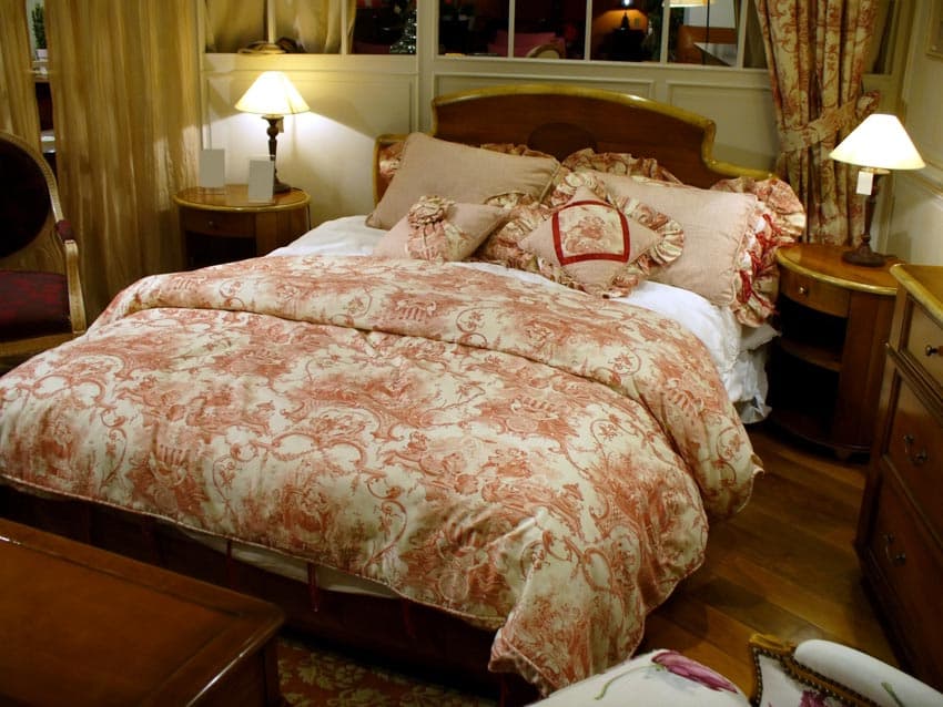 Bedroom with thick comforter, nightstands, lamps, pillows, wood flooring, and window curtain