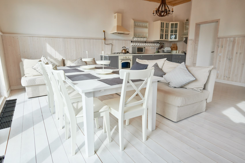 Bright dining room with white floors, table, chairs, kitchen area, and wainscoting