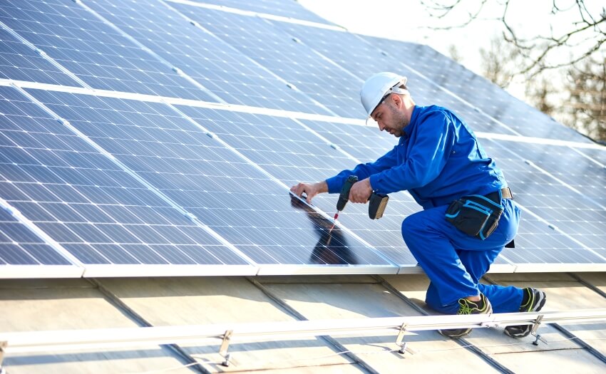 Electrician in blue suit installing solar panel on roof of modern house