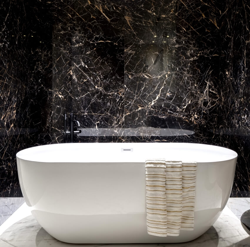 Freestanding bathtub in elegant bathroom with black marble tiles on walls and beige marble floor