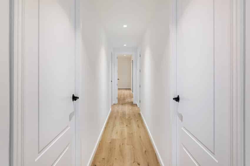 Interior of long narrow hallway with closed white doors, wooden floor, and white walls