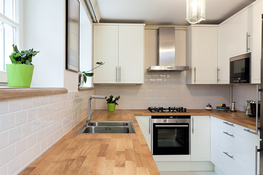 Kitchen with wood tile countertop, white cabinets, tile backsplash, range hood, stove, and oven