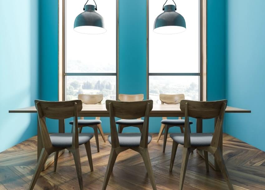 Narrow dining area with blue walls, windows, wooden table with brown seats, and hanging lights