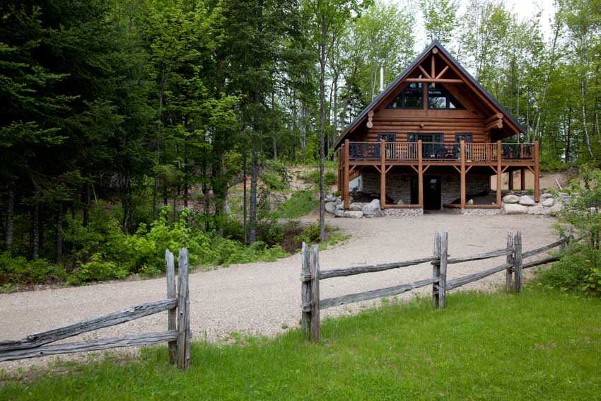 Road leading to modern log cabin with elevated deck, windows, and roof
