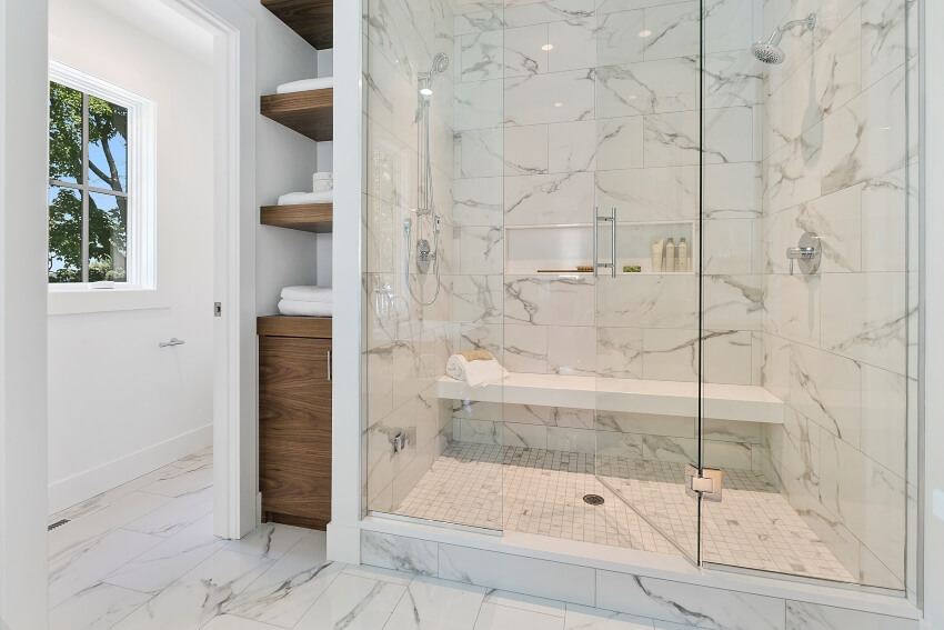 Shower enclosed in glass with marble tile wall and bench, and wood shelves on the side