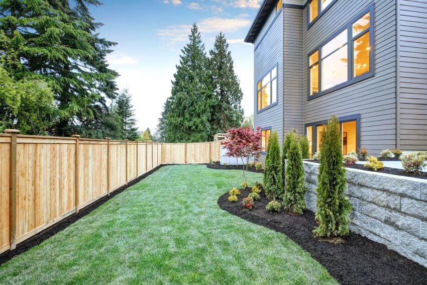 Three-story wood siding home exterior with manicured lawn, flower beds, and cedar wood fence