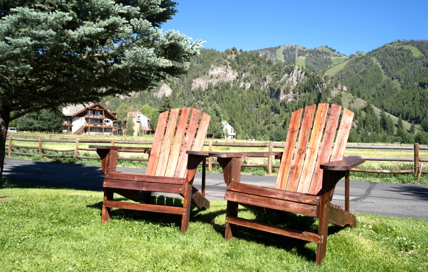 Two empty wood adirondack chairs sit on grass overlooking the mountains