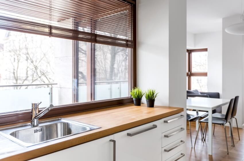 White kitchen with wood countertop, silver sink, big window with blinds, and view of the dining area