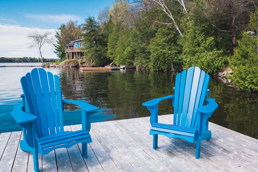 A pair of blue Muskoka chairs on a deck beside a lake