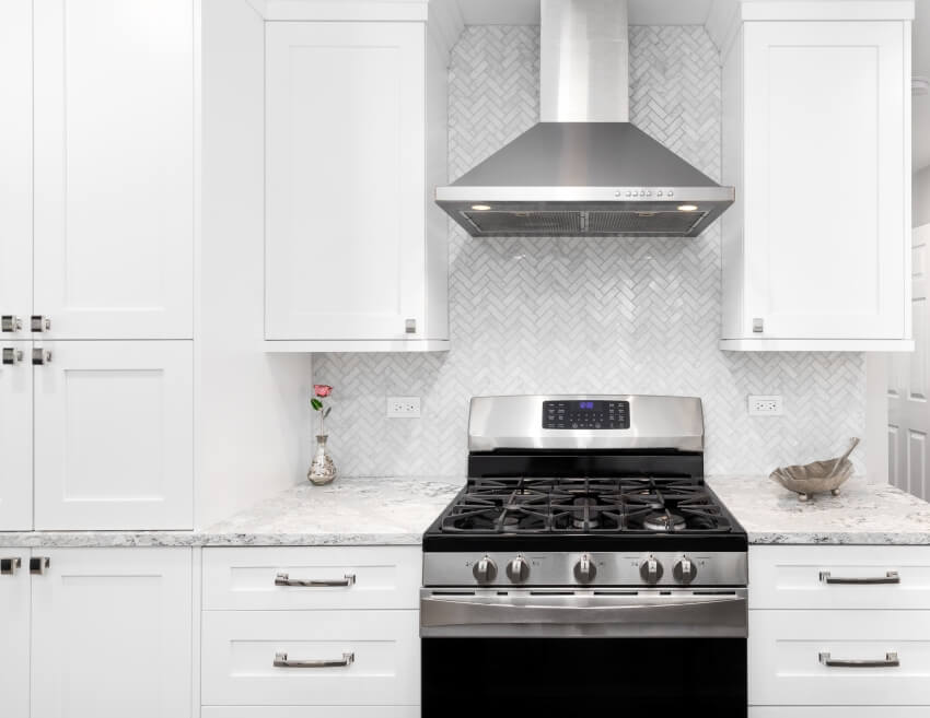 A stainless steel oven and hood in a white kitchen with calcatta marble countertops and a herringbone backsplash