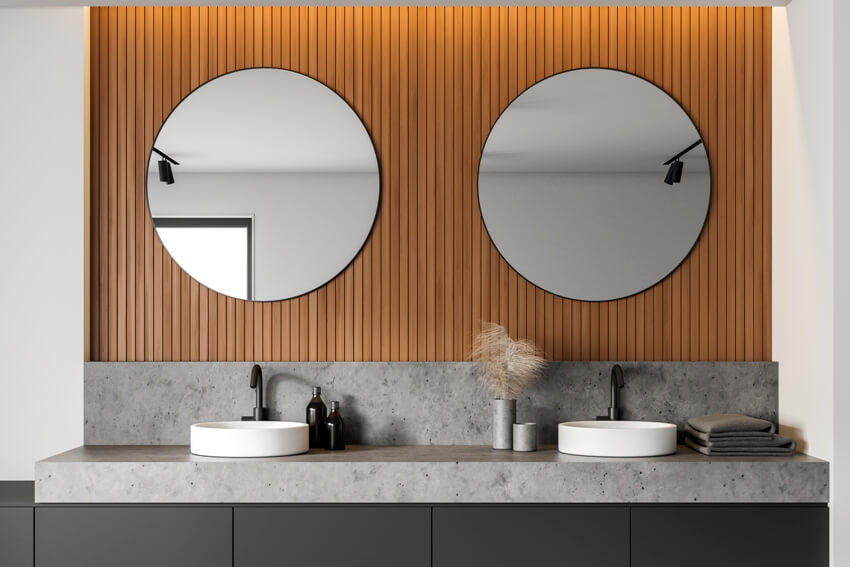 A close up of double bathroom sink on concrete laminate countertop in stylish room with white and wooden walls and two round mirrors