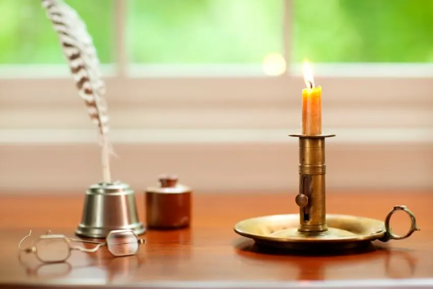 A colonial candle in chamberstick quill pen and glasses on desk with window background