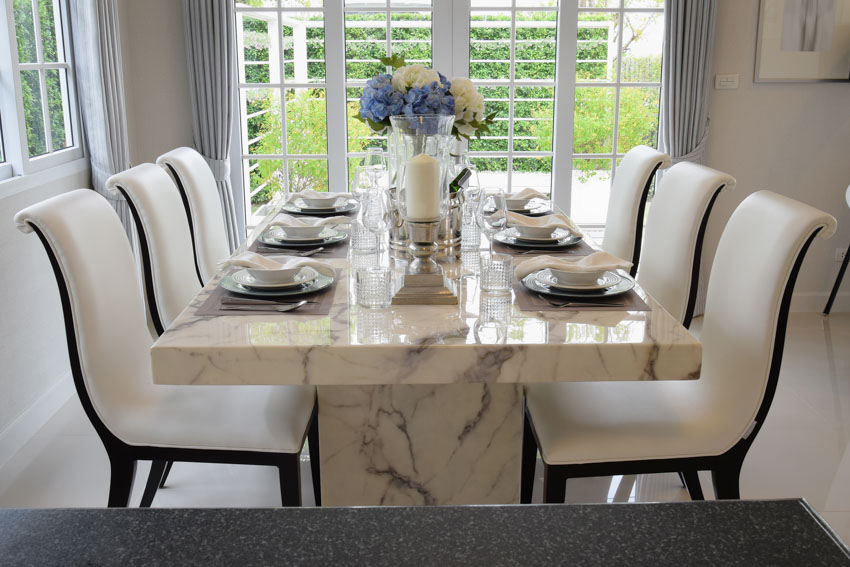 Dining room with marble table, white chairs, plates, vase, flowers, and windows