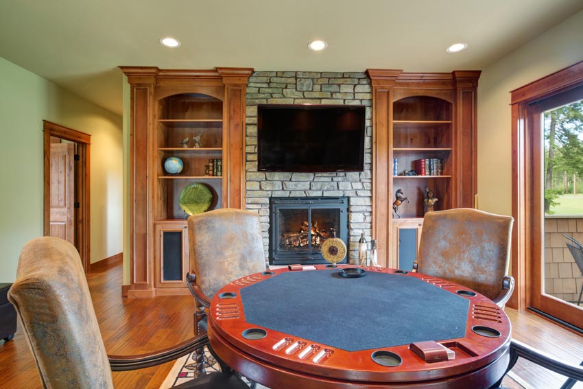 Living room with card table, brick fireplace, wood shelves, chairs, wooden flooring, and glass window
