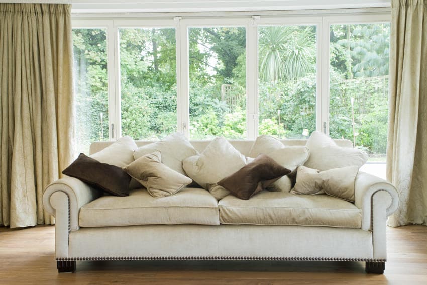 Living room with cushioned white couch, pillows, wood floor, curtains, and triple pane windows