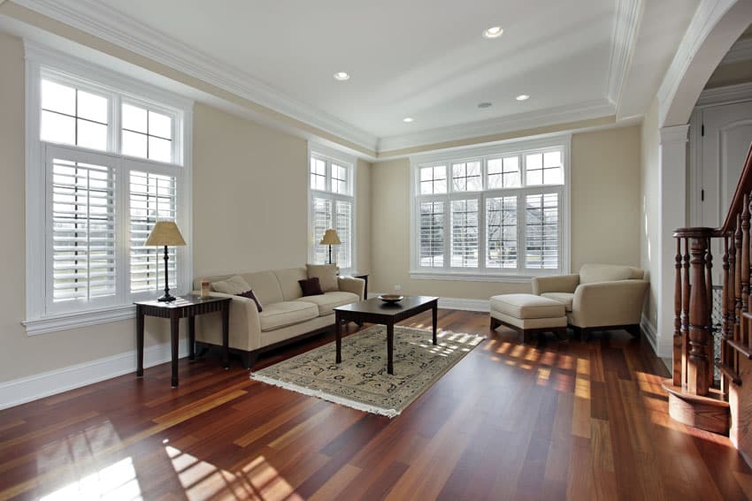 Room with plantation style windows, white walls, wood floors, couch, sofa chair, hassock, and coffee table