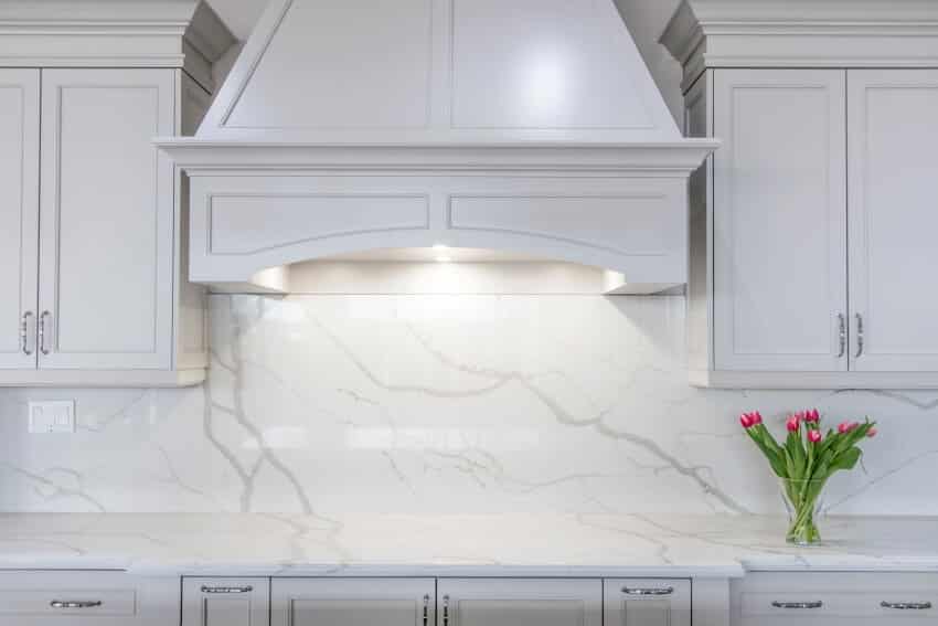 A modern white kitchen with a traditional touch featuring white calacatta marble countertop and backsplash, white cabinetry and custom made range hood