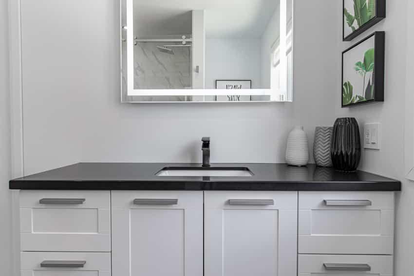 Simple bathroom vanity with mirror, sink, faucet, black laminate countertop, and white cabinets