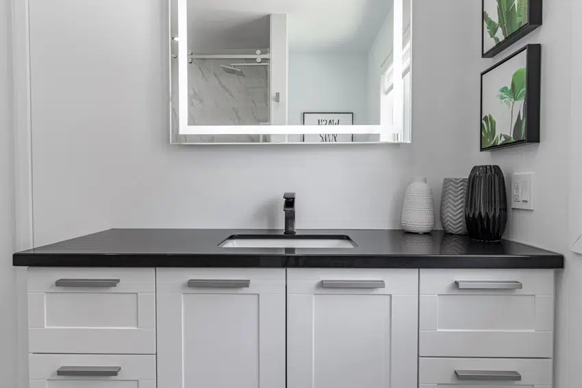 Simple bathroom vanity with mirror, sink, faucet, black laminate countertop, and white cabinets
