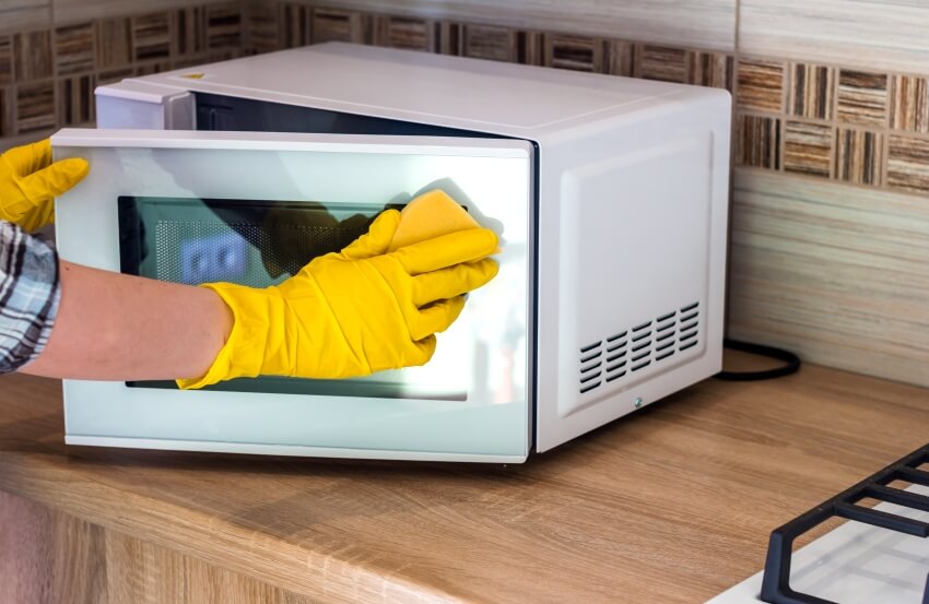 A woman's hands in yellow gloves wiping microwave with sponge