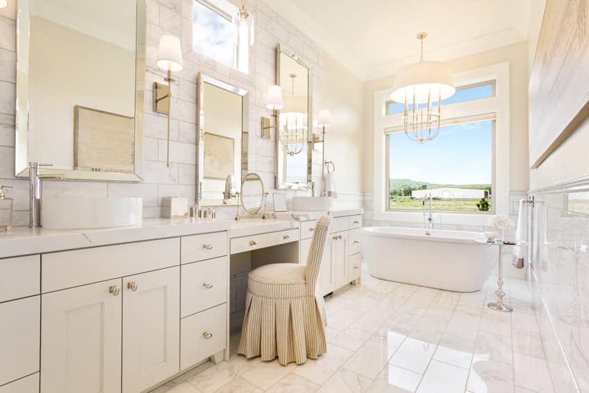 Bathroom with floor to ceiling tiles, chair, vanity area, window, tub, hanging light, wall mounted lights, and window