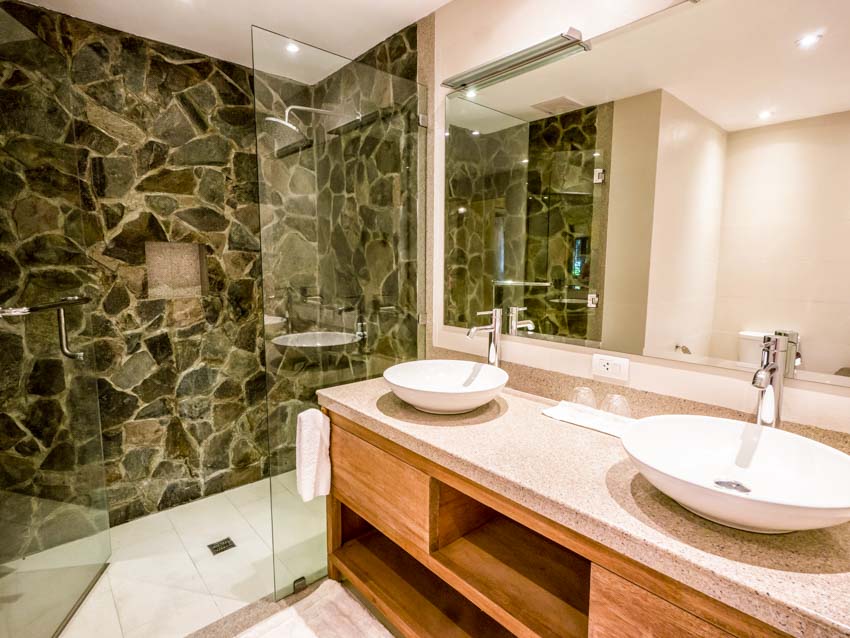 Bathroom with stone shower wall, glass divider, granite countertop, basin sinks, faucet, and mirror
