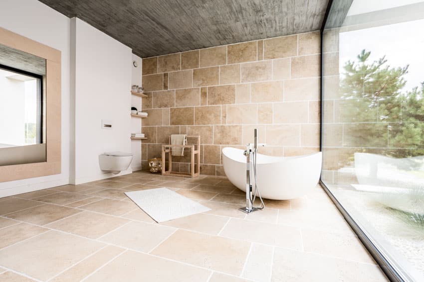 Bathroom with textured ceramic floor to ceiling tiles, tub, toilet, floating shelves, and window