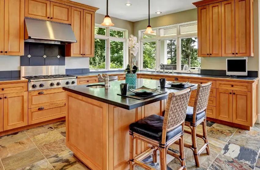 A bright spacious kitchen interior with steel appliances, wooden cabinets, and island with bluestone countertop and chairs