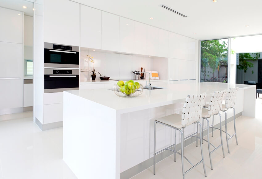 Clean pure white kitchen interior with built in microwave and oven, long island with sink and chairs and white cabinets
