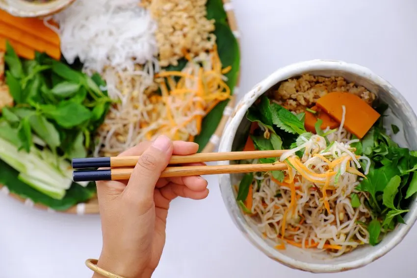 A hand with chopsticks getting some in a bowl of vegan noodles with vegetables and fresh oregano