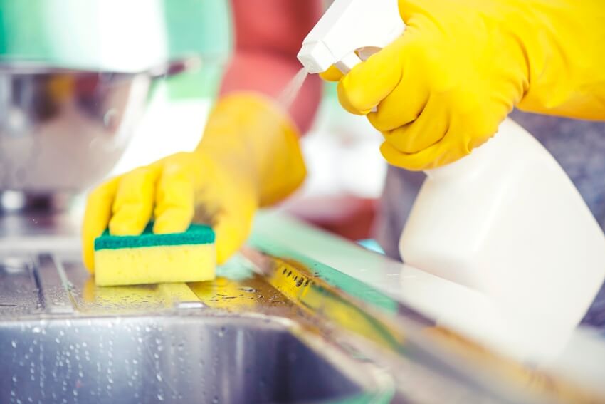Hands with yellow gloves cleaning a kitchen sink with sponge and spray detergent