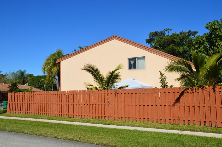 House exterior with palm trees, beige painted wall, pitched roof, and board on board wood fence