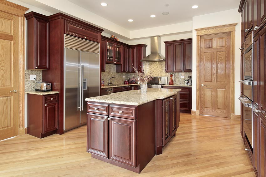 Kitchen with light wood floors, mahogany cabinets, center island, countertops, backsplash, range hood, refrigerator, and ceiling lights