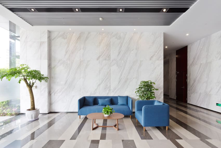 Living room with bookmatched porcelain wall, blue couch, accent chair, coffee table, indoor plants, and ceiling lights