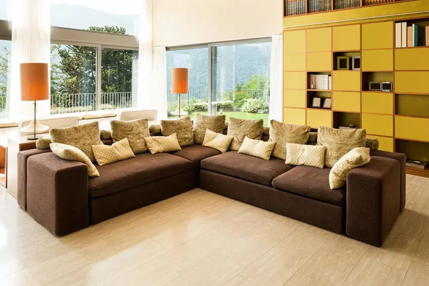 Living room with brown cushioned couch, pillows, birch wood floors, lamp, shelves, windows, and curtain