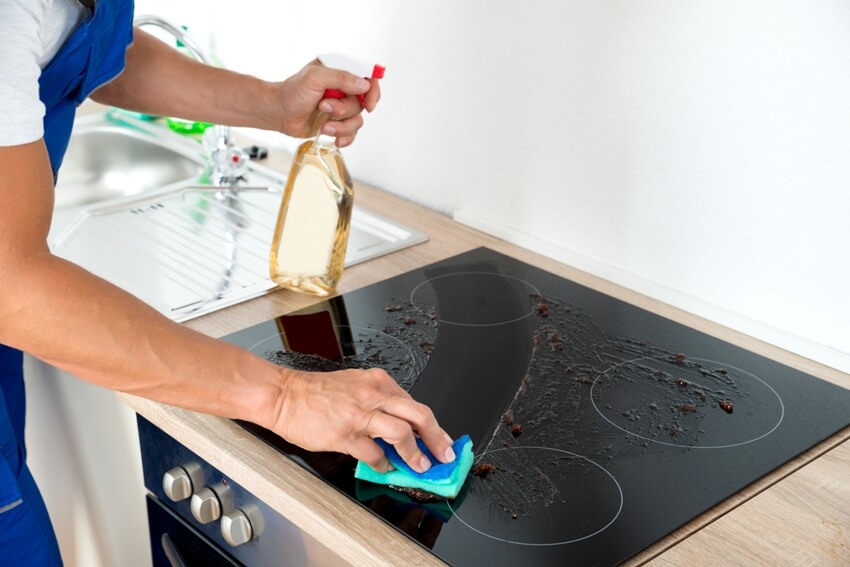 A man cleaning a messy induction stove with detergent spray and sponge in kitchen