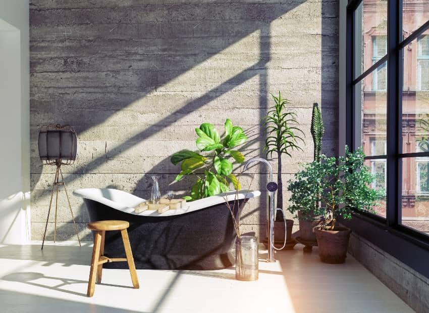 A modern industrial design bathroom with solid concrete wall accent, freestanding bathtub, natural lighting, and big windows