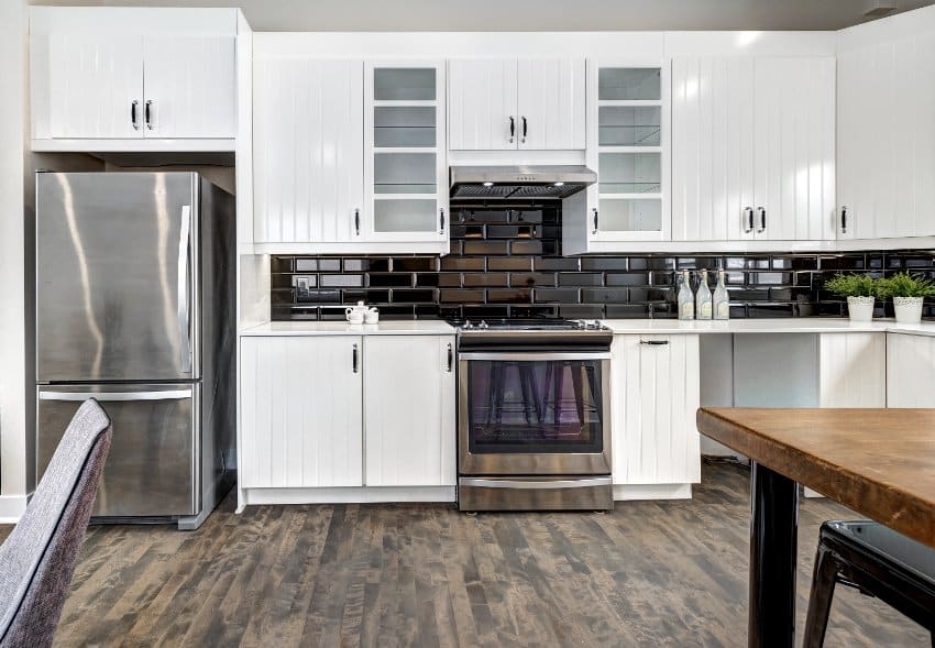 Ktchen interior with wood tile flooring, white grooved cabinets, black subway tile backsplash and stainless steel appliances