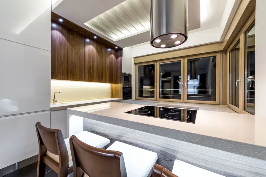 Modern kitchen with countertop, backsplash, walnut cabinets, range hood, induction stove, ceiling lights, and windows