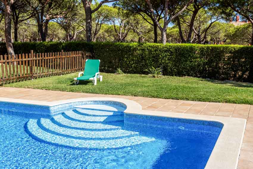Outdoor swimming pool with fence, lounge chair, hedge plants, and glass bead finish