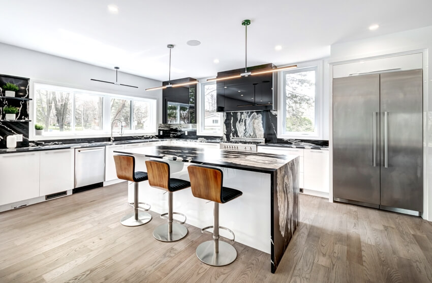 Spacious and clean kitchen interior with waterfall marble island counter, stainless steel fridge, wooden floors and black and white cabinets