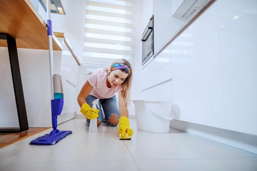 A woman with rubber gloves kneeling in kitchen and cleaning kitchen floor with sponge and sprayer