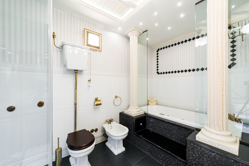 Bathroom with stepped tub, black and gold finishes, toilet, tub, white paneling, and pillars