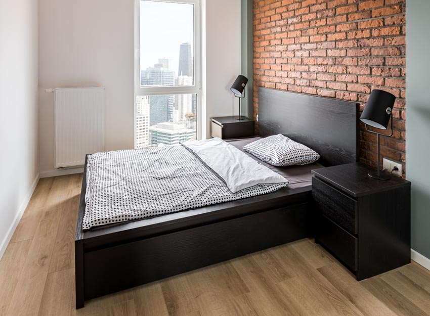 Bedroom with brick wall cladding, bed, headboard, nightstands, lamps, and wood floors
