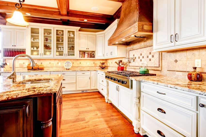 Classic kitchen with range hood, white cabinets, wood floors, sink, faucet, countertops, and limestone backsplash