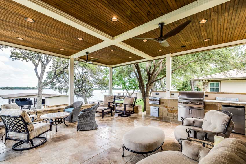 Covered patio with travertine pavers, natural stone outdoor kitchen, table, chairs, wood slat ceiling, and lighting fixtures