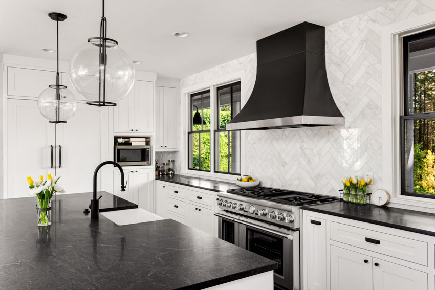 Kitchen with black granite countertop, marble herringbone backsplash, range hood, island, pendant lights, and windows